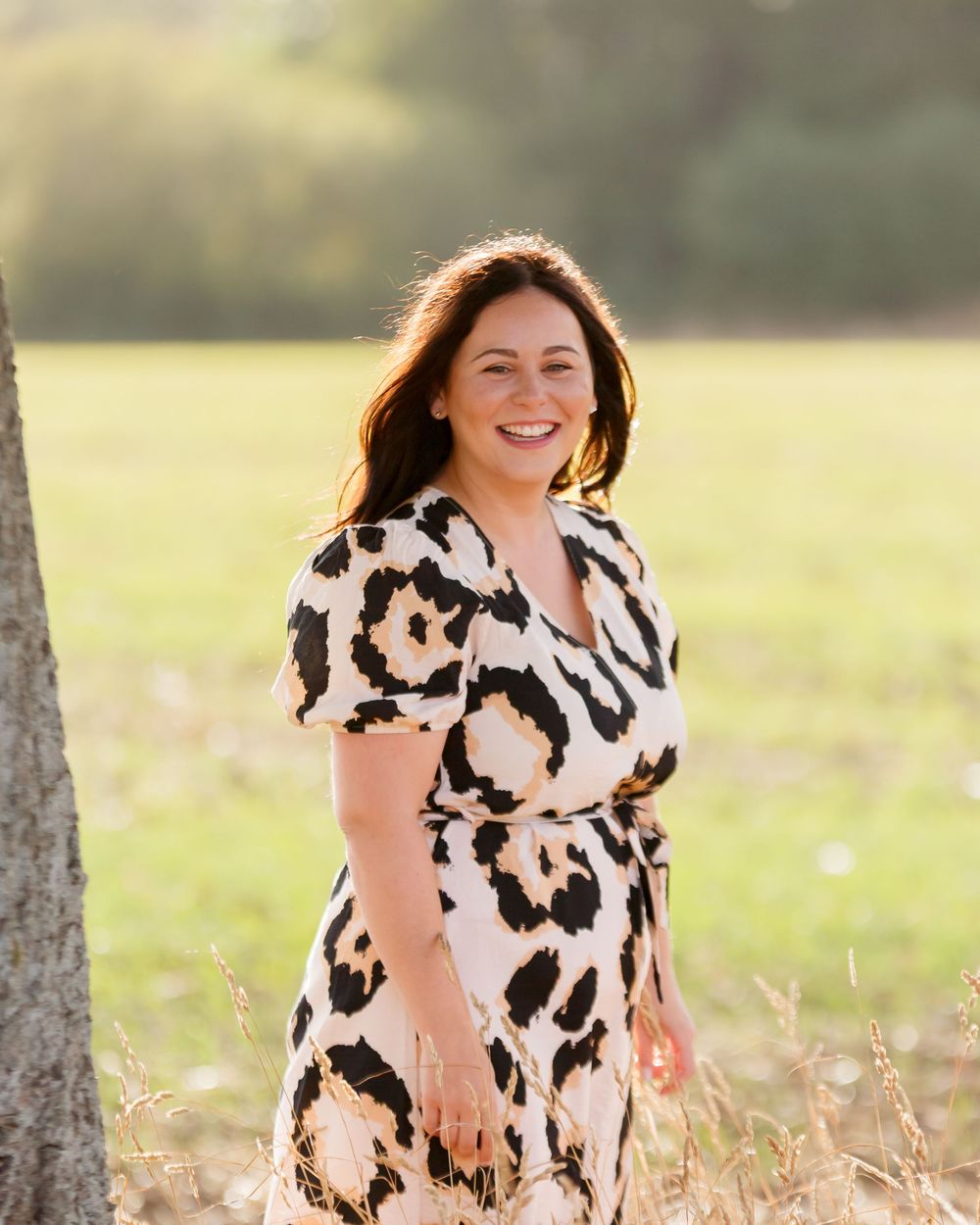 A woman in a spotted dress sits joyfully in tall grass during a sunny outdoor photo session.