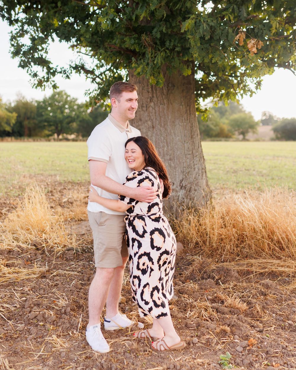 Man and woman share a playful moment beneath a large tree in a sunlit field during autumn.