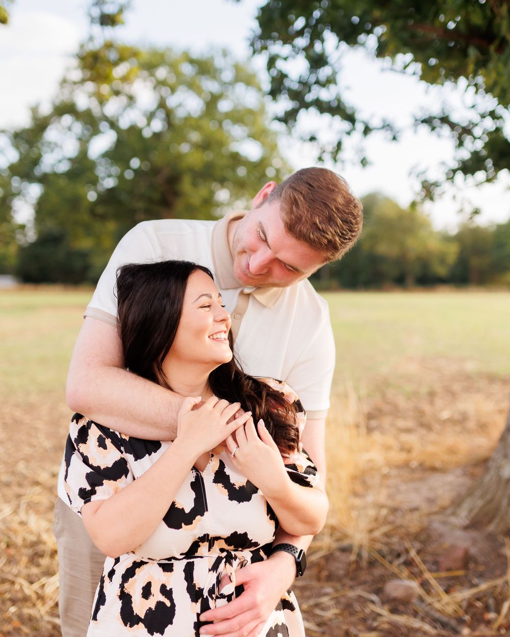 Romantic couple embraces in a sunlit field with golden grass and trees in the background.