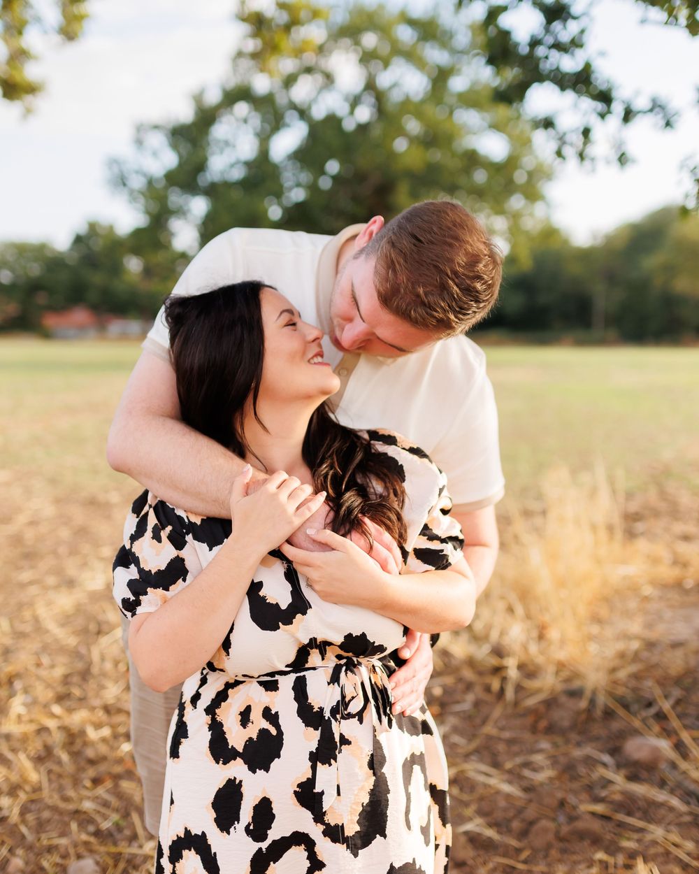 A couple shares a tender embrace in a golden field during sunset, with her wearing a black and white patterned dress.