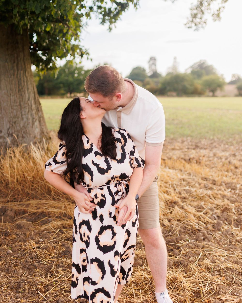 A romantic moment captured between a couple standing by a tree in a summer field with warm lighting.