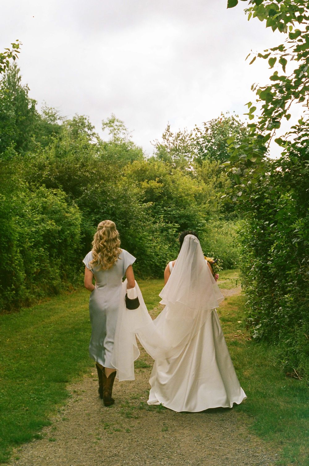 Wedding party walking down a tree-lined path holding up the bride's dress train.