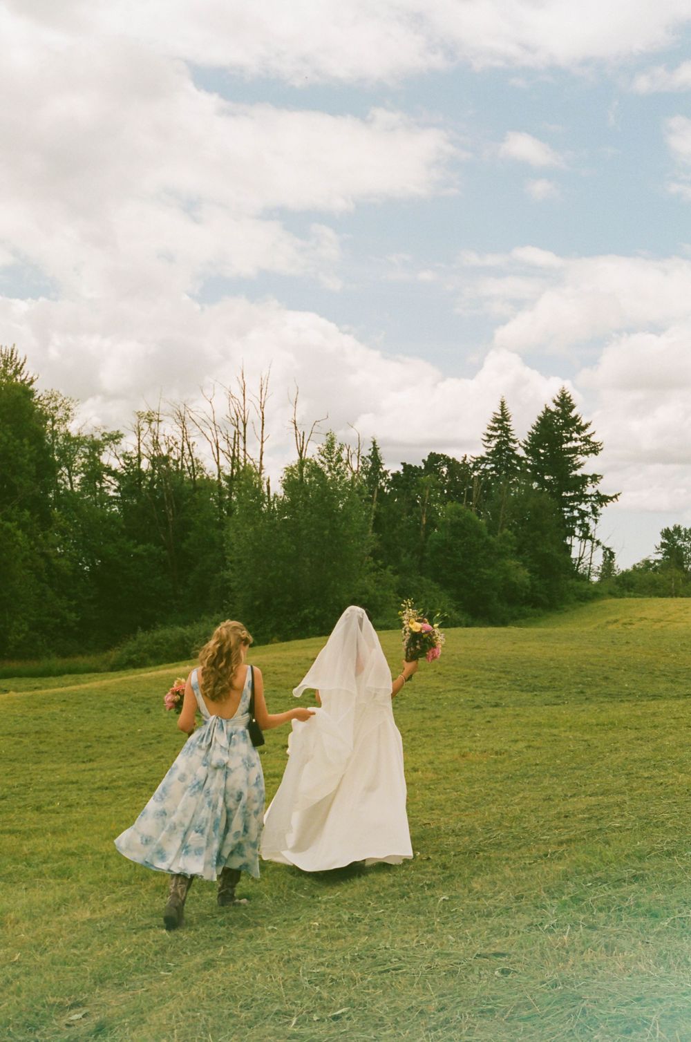 Two figures in floral dress and white veil run playfully across a grassy hillside on a cloudy summer day.
