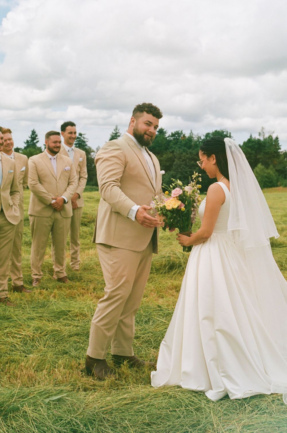 Wedding party in tan suits and white dress pose together in an outdoor meadow setting.