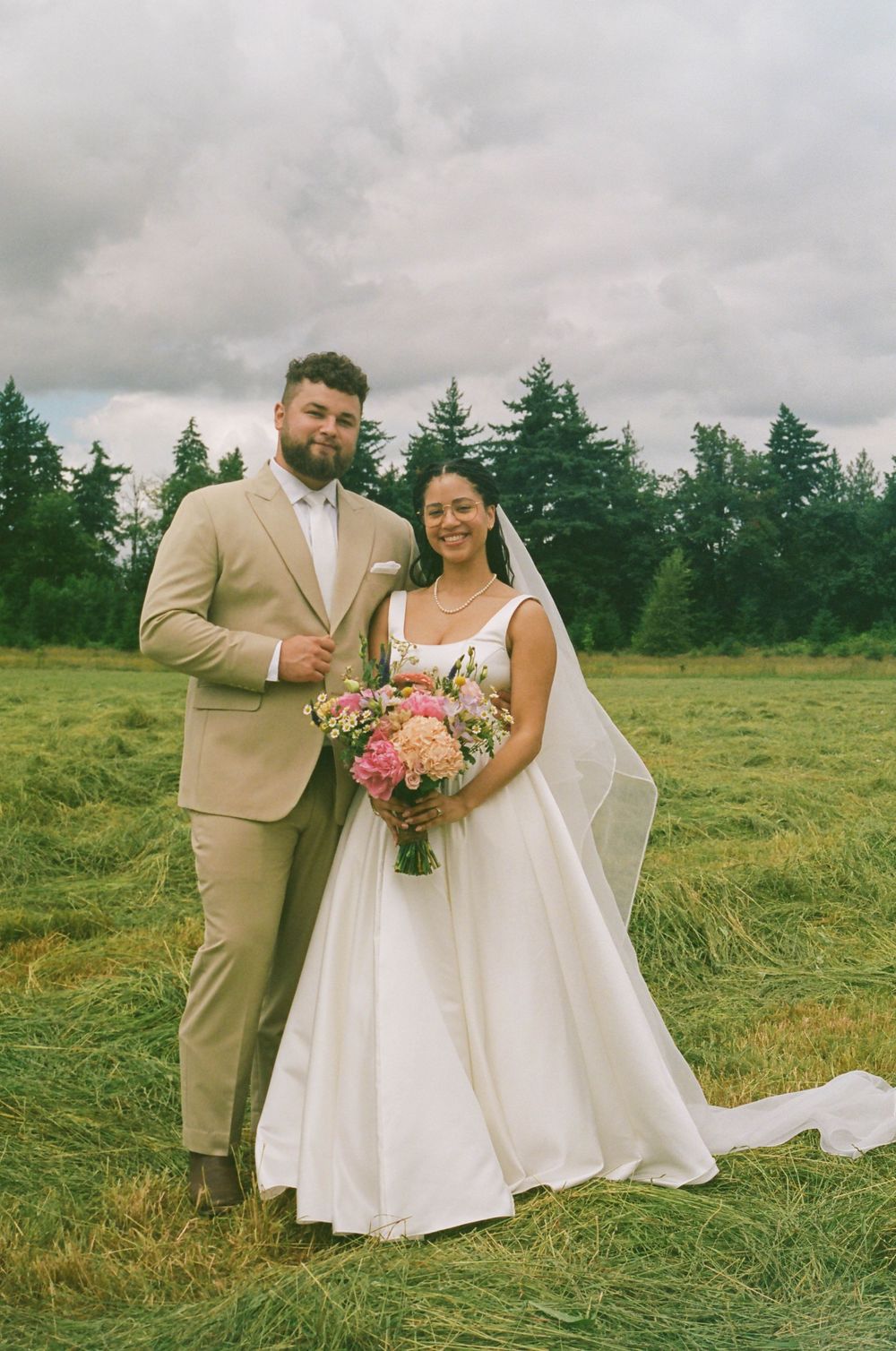 Wedding couple poses in tan suit and white dress holding pink bouquet in grassy field.