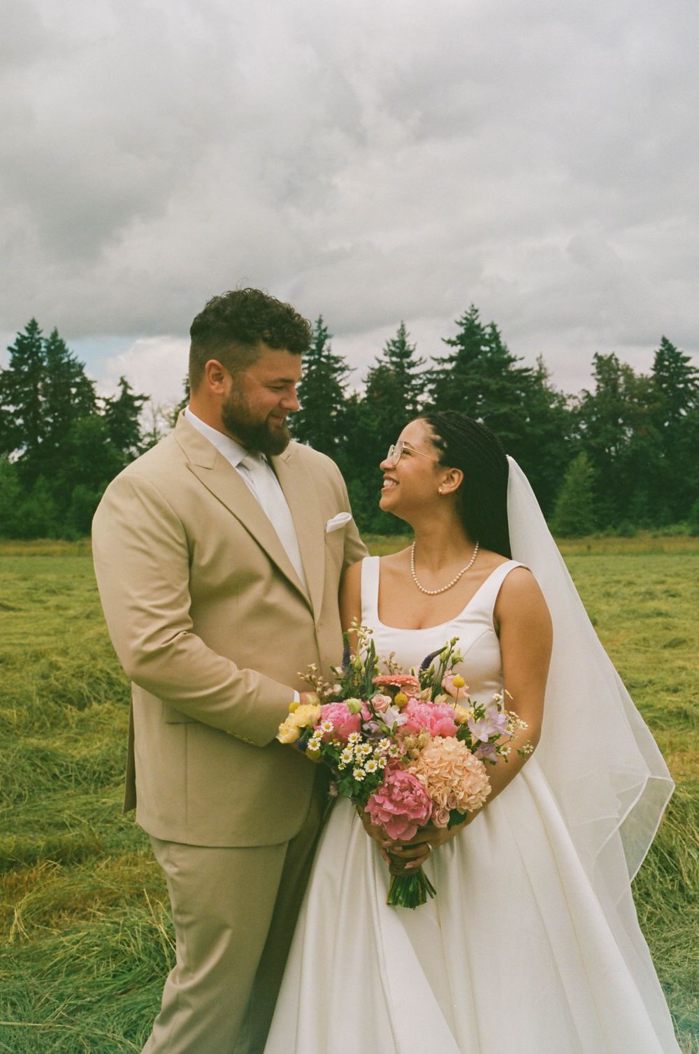 Happy wedding couple laughing together holding pink floral bouquet in outdoor setting.