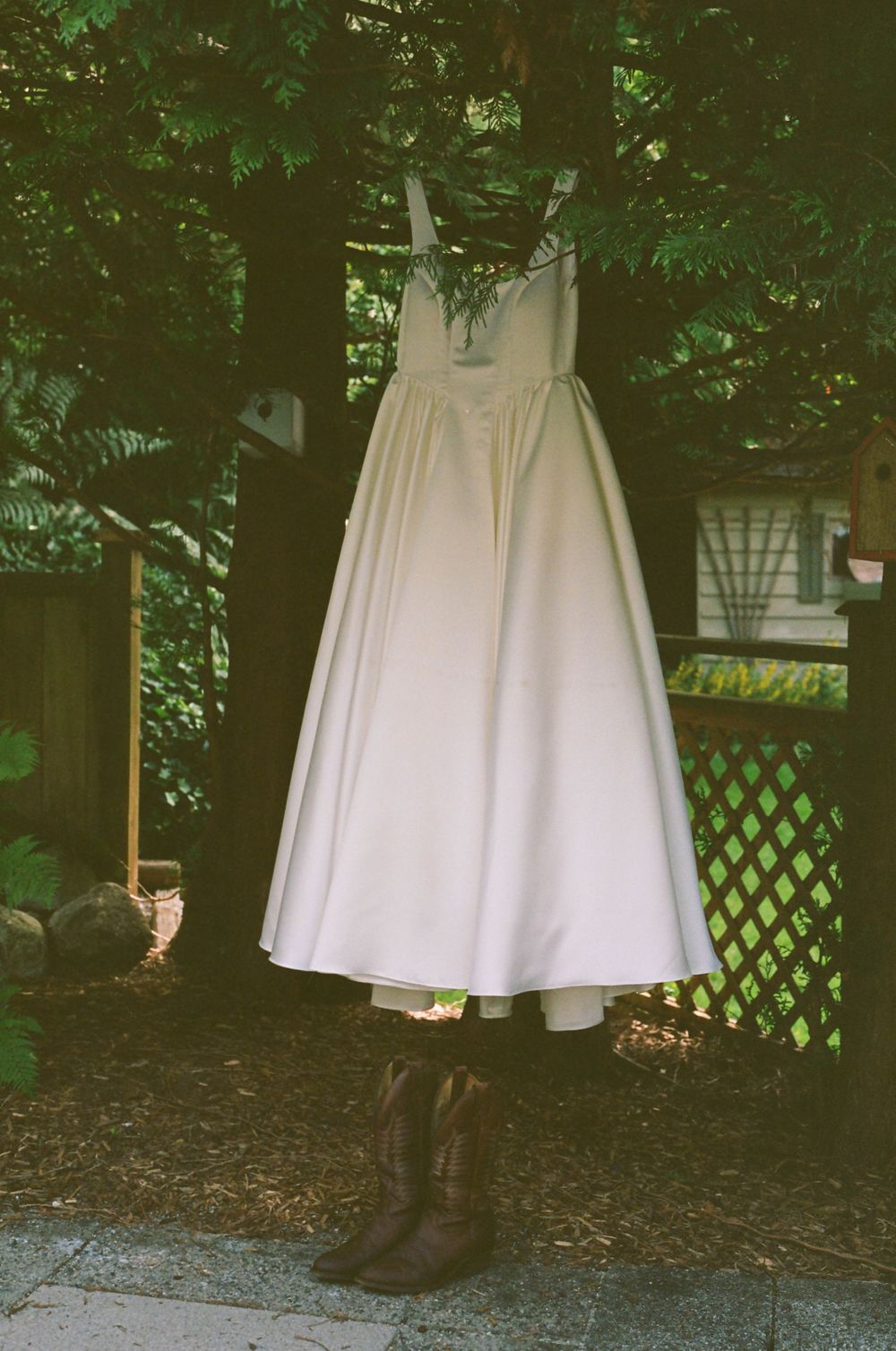 White wedding dress photographed hanging outdoors against green foliage and lattice backdrop.