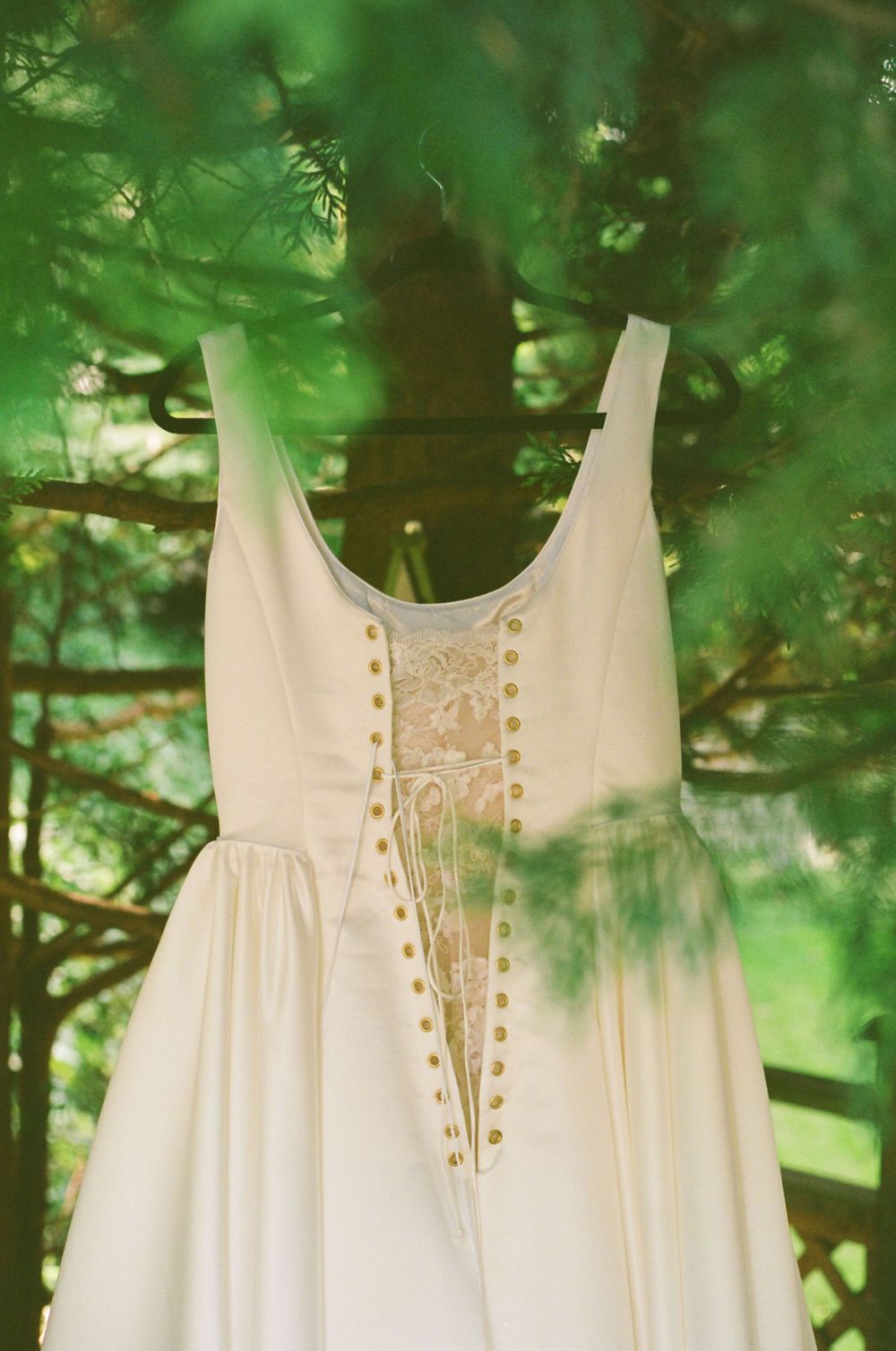 Close-up detail of white wedding dress with decorative buttons and lace pattern against green foliage.