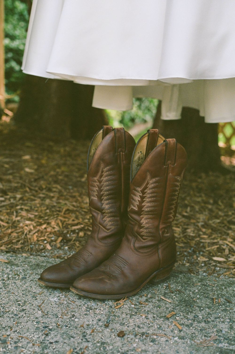 White wedding dress hanging above brown leather cowboy boots on gravel ground.