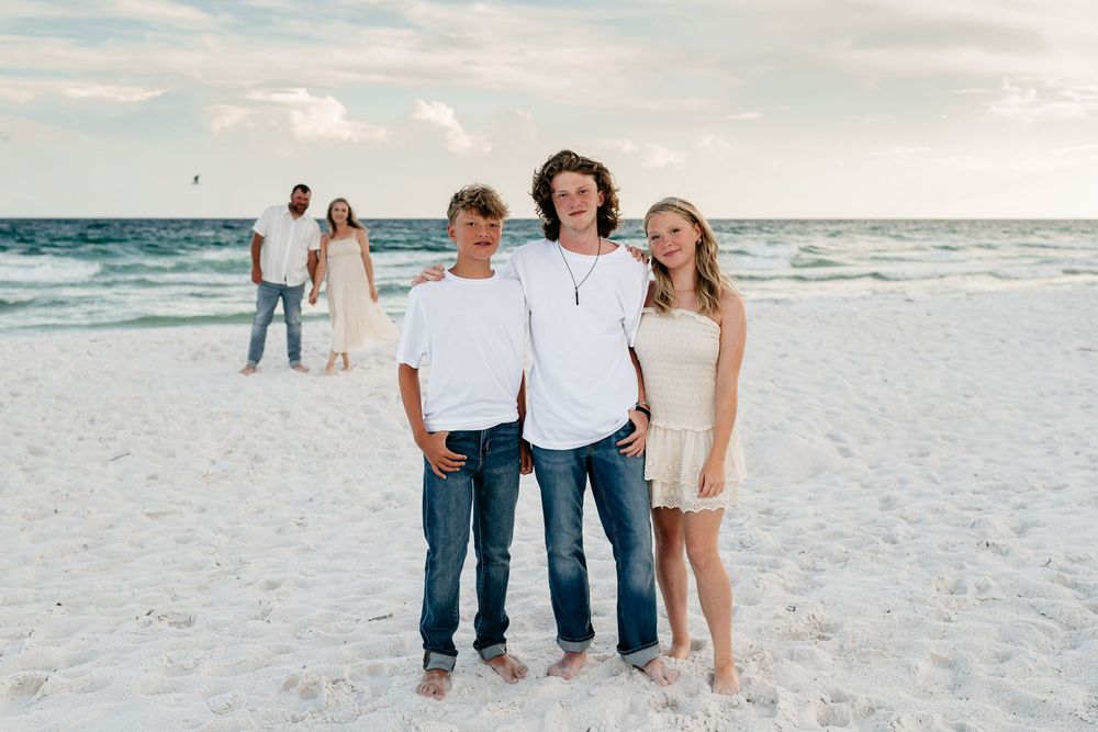 Family members wearing jeans and white shirts walk together on a pristine white sandy beach at sunset.