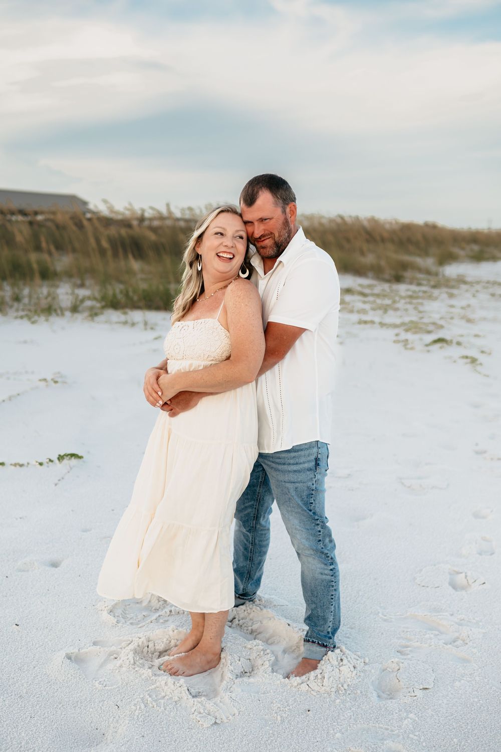 A couple embraces joyfully on a pristine white sand beach with sand dunes and sea grass in the background.