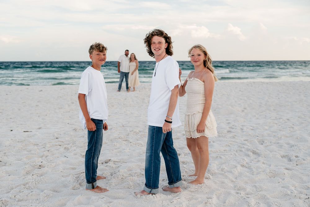 Family in white clothing poses together on a sunny white sand beach during sunset near the ocean.