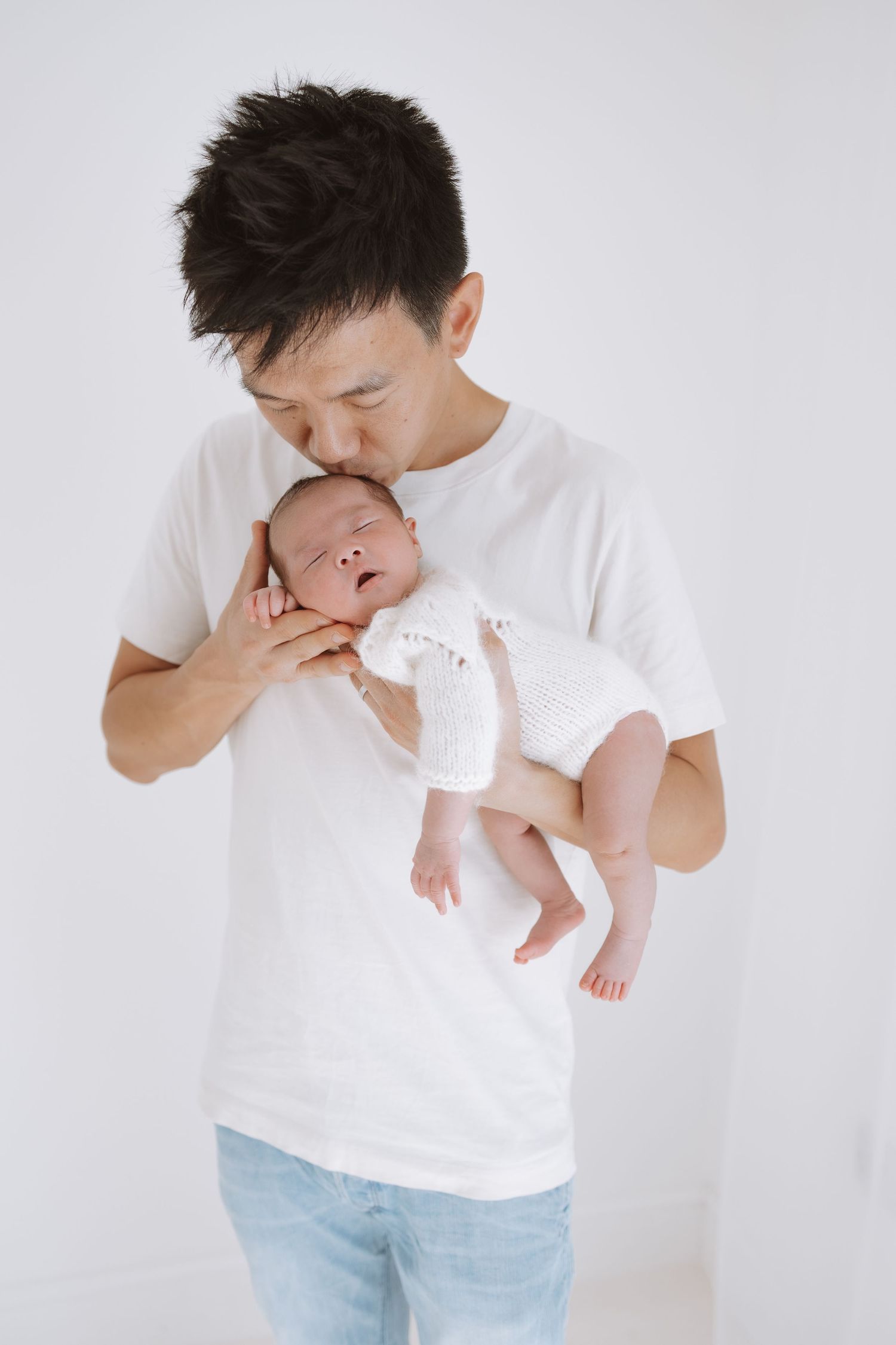 Person in white t-shirt and jeans tenderly holding and kissing a sleeping newborn baby wearing white.