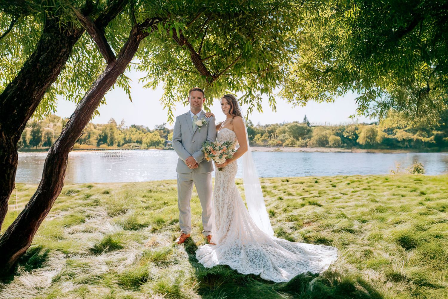Elegant couple in wedding attire poses under a willow tree beside a serene lake on a sunny afternoon.