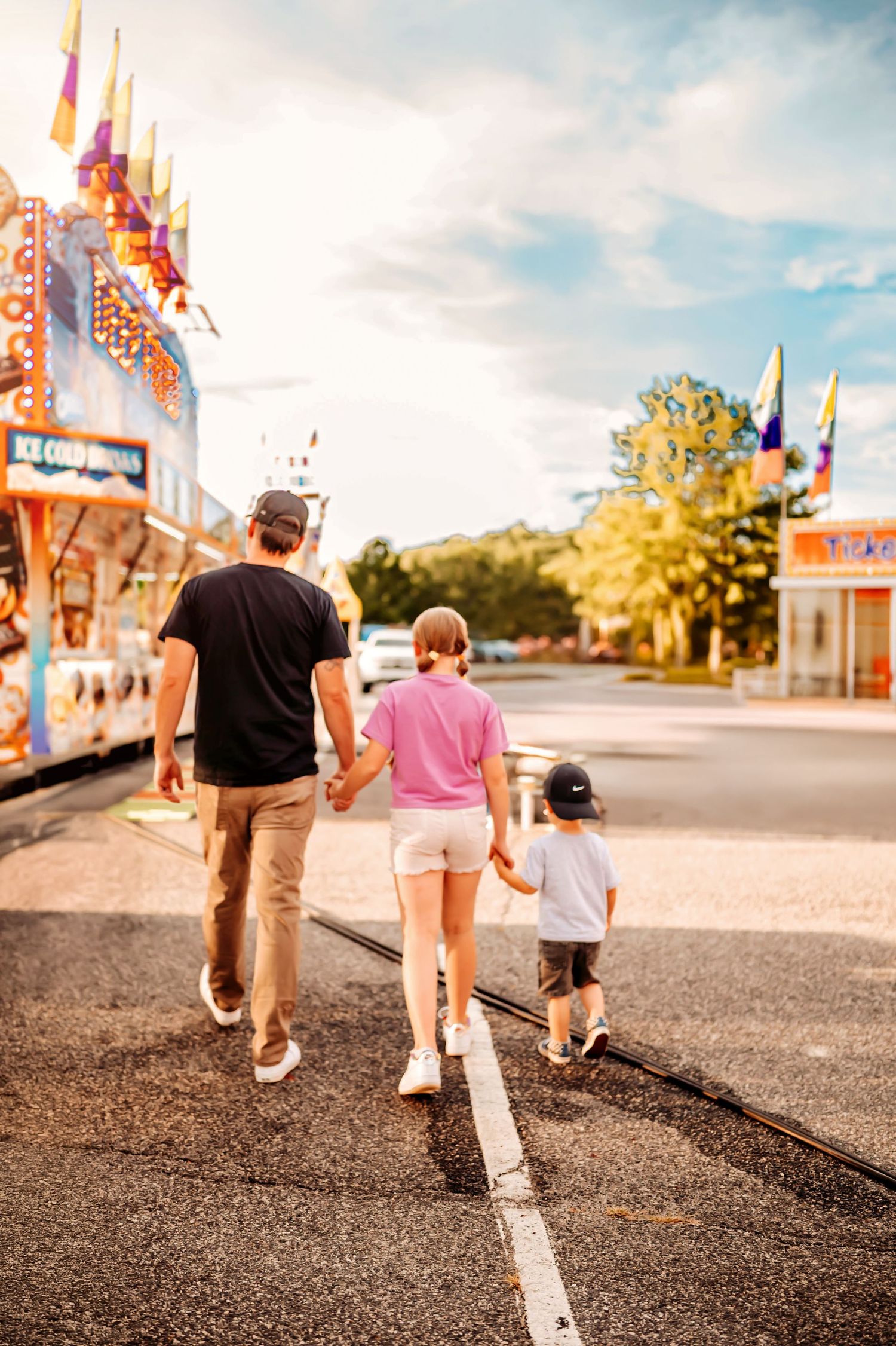 A family walks together down a carnival midway on a sunny day with game booths and colorful attractions visible.