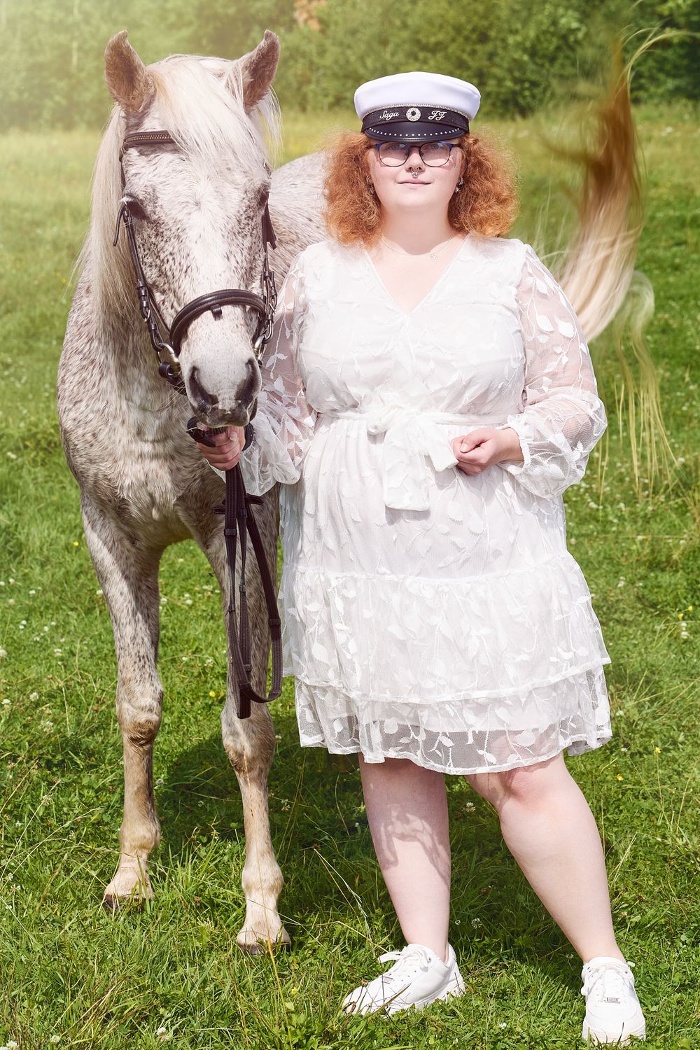 A white horse stands next to a person wearing a white lace dress and graduation cap in a sunny meadow.