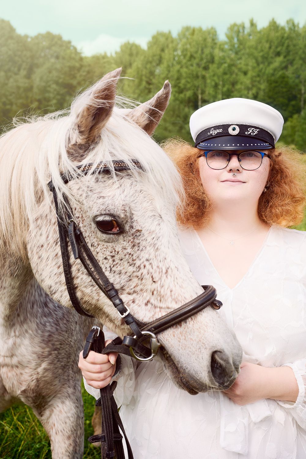A dappled gray horse stands next to someone wearing a white naval cap and white clothing outdoors.