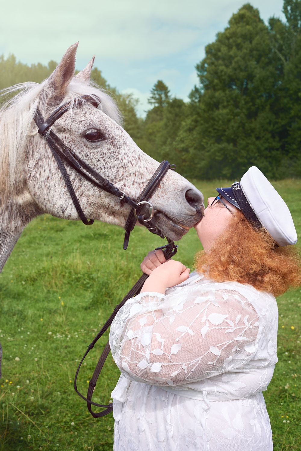 A dappled gray horse playfully interacts with a person wearing a white dress and sailor hat in a pastoral setting.