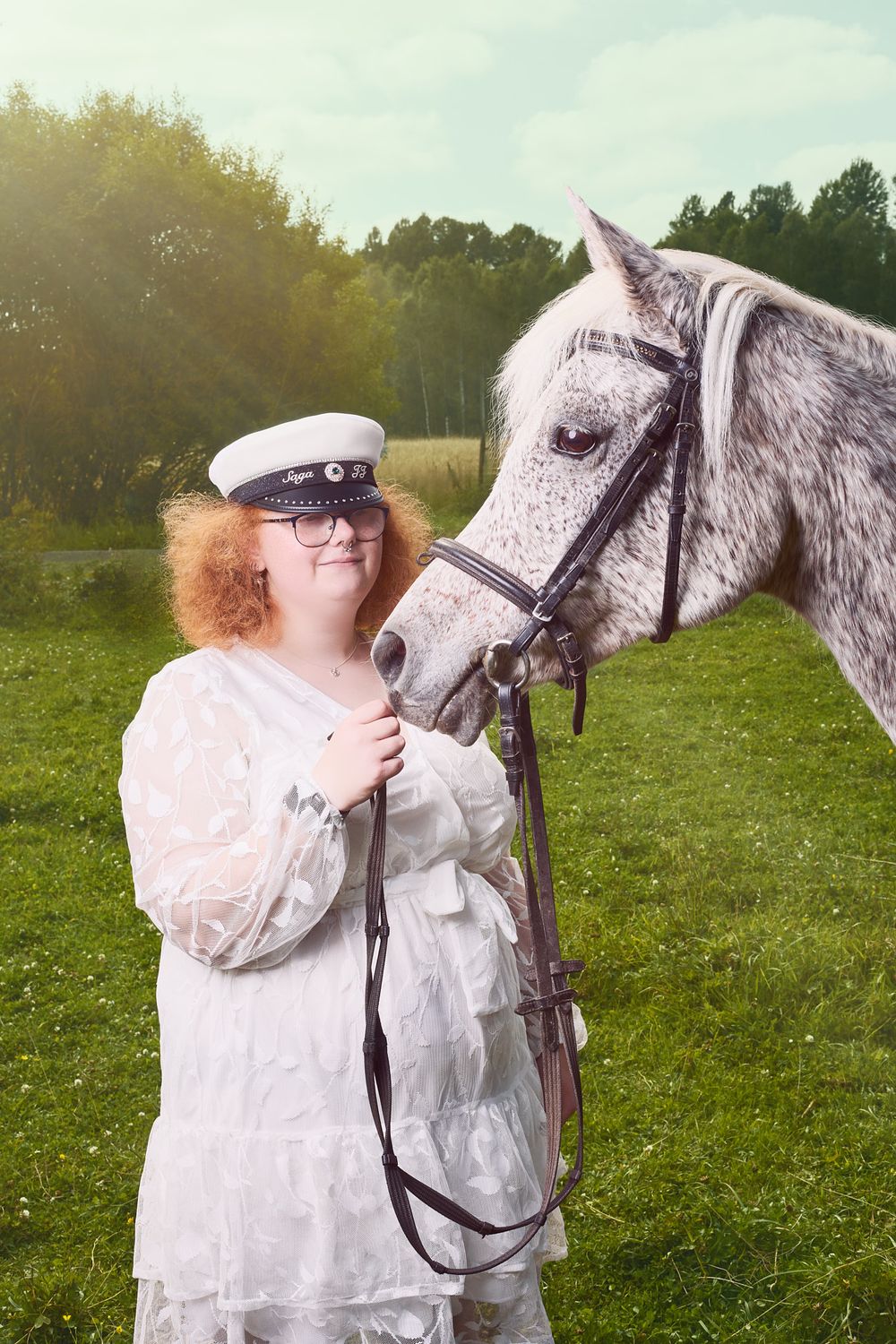 A dappled gray horse stands beside a person in a white dress and sailor cap on a grassy field.