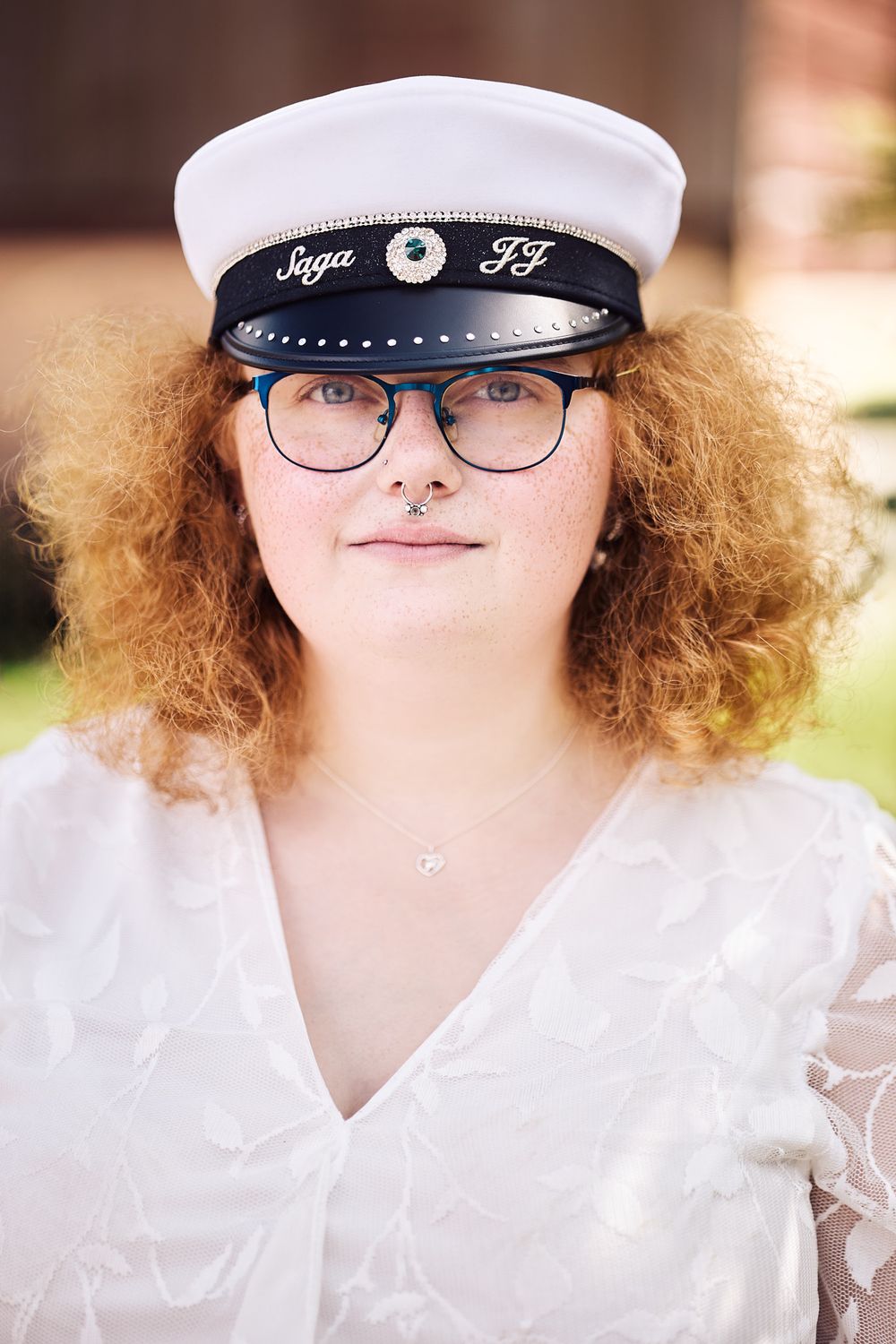 Graduation portrait showing white cap with "Saga FF" emblem, curly ginger hair, glasses, and white lace dress.