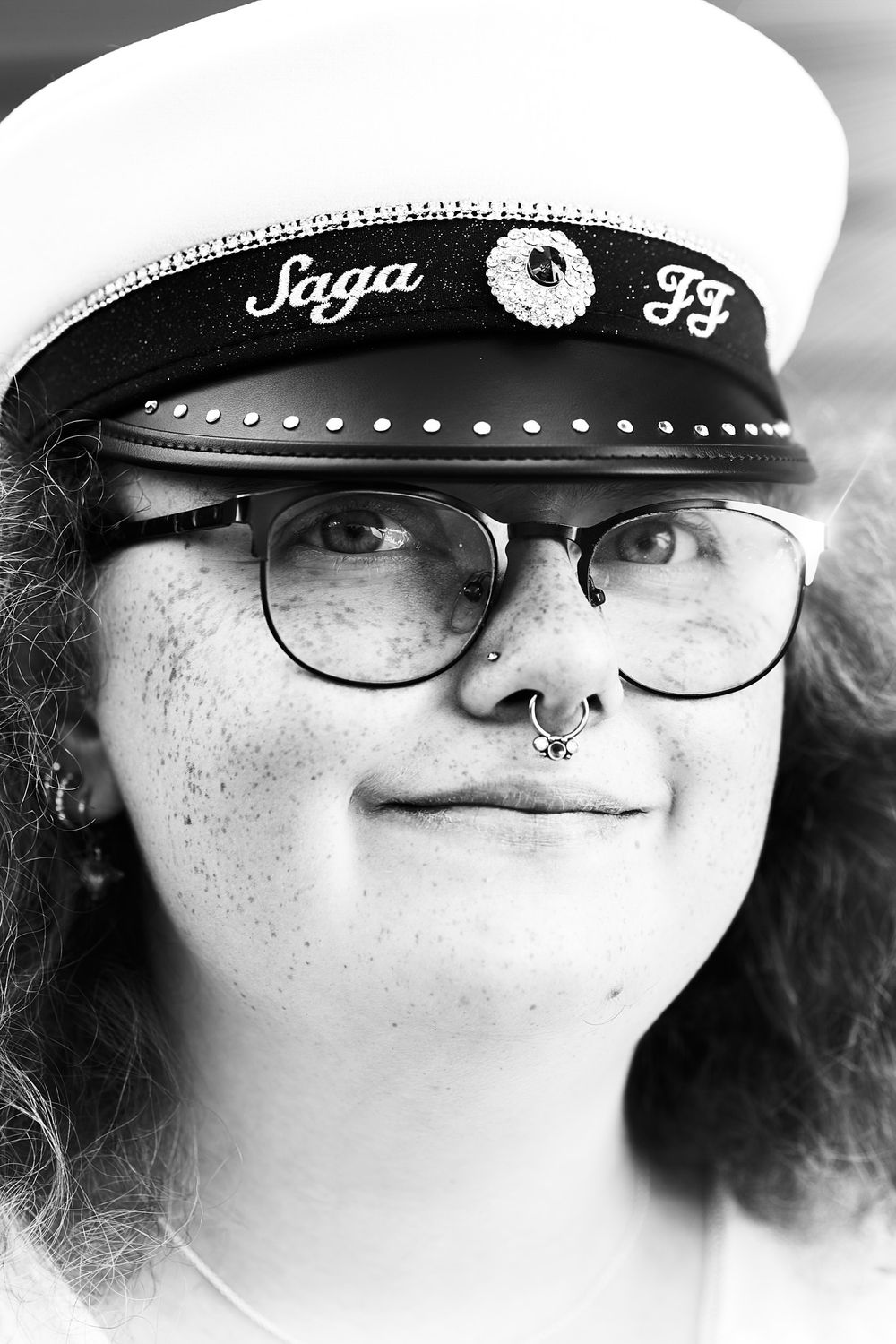 Black and white portrait with student cap labeled "Saga FF", glasses, and a septum piercing.