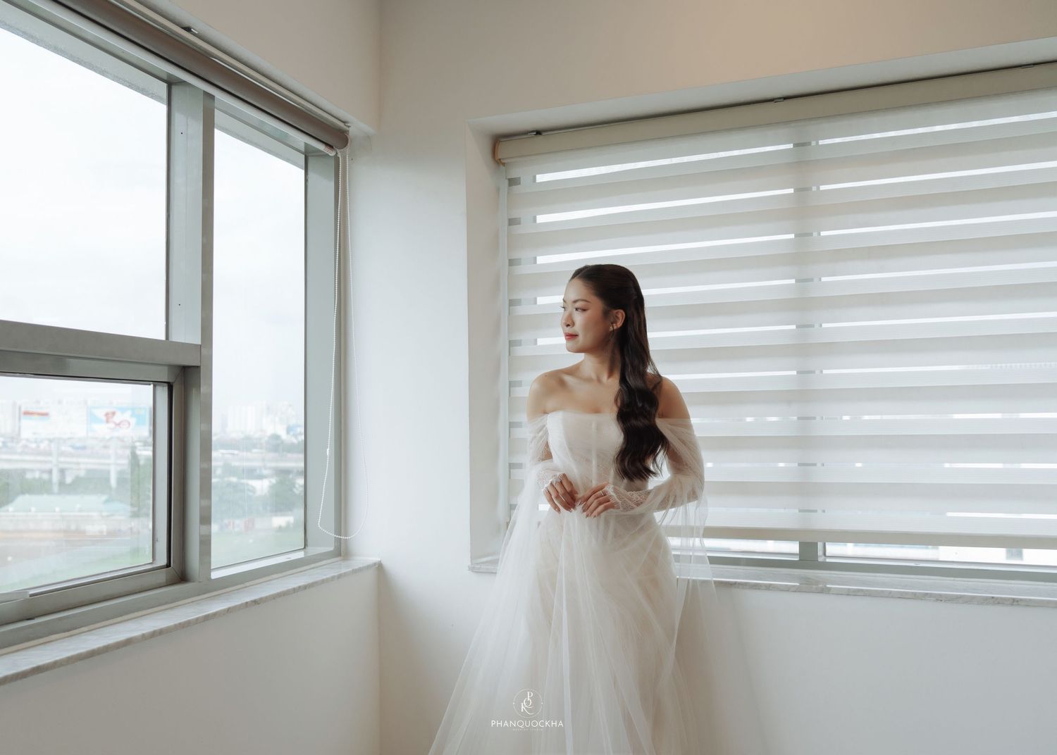 Bride in elegant off-shoulder white tulle wedding gown standing by large windows in soft natural light.