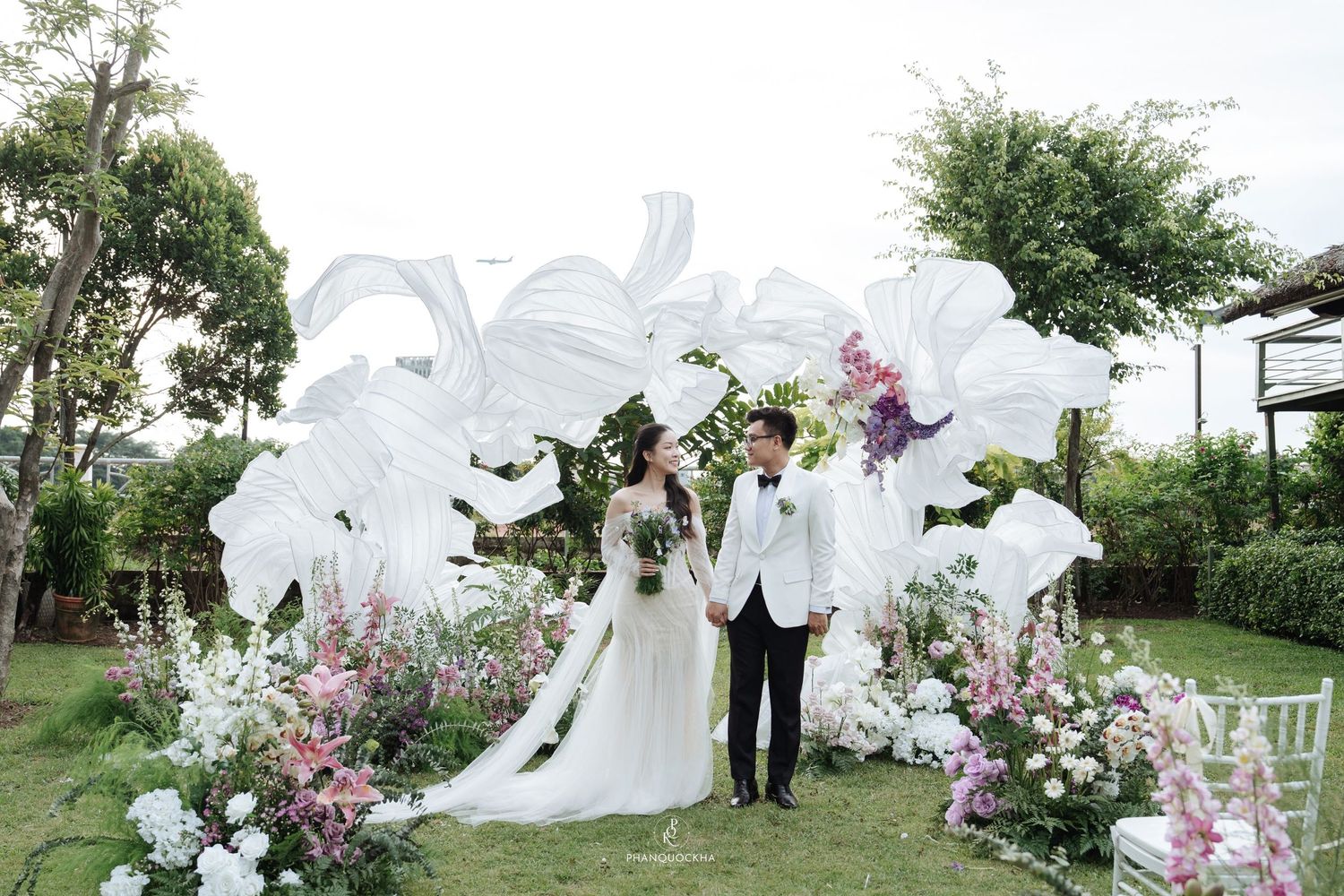 Bride and groom hold hands before stunning white fabric floral arch surrounded by pink and white flowers outdoors.