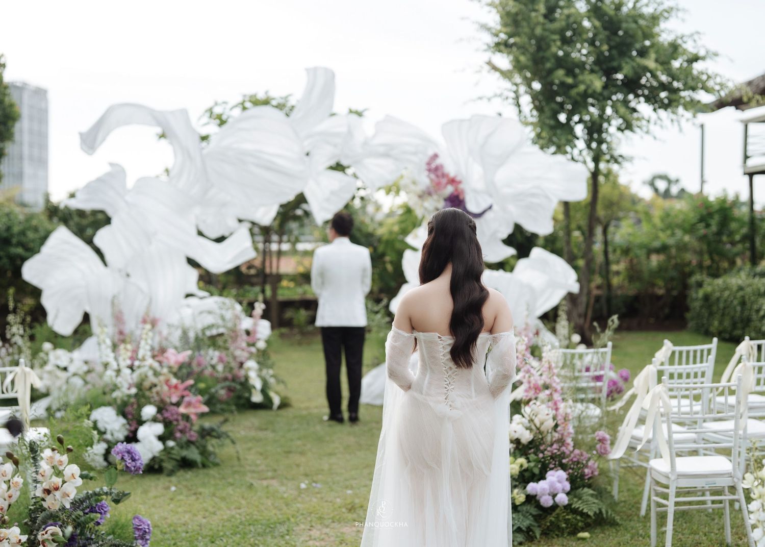 Bride in white gown walks toward floral arch with large white fabric sculptures at outdoor garden wedding ceremony.