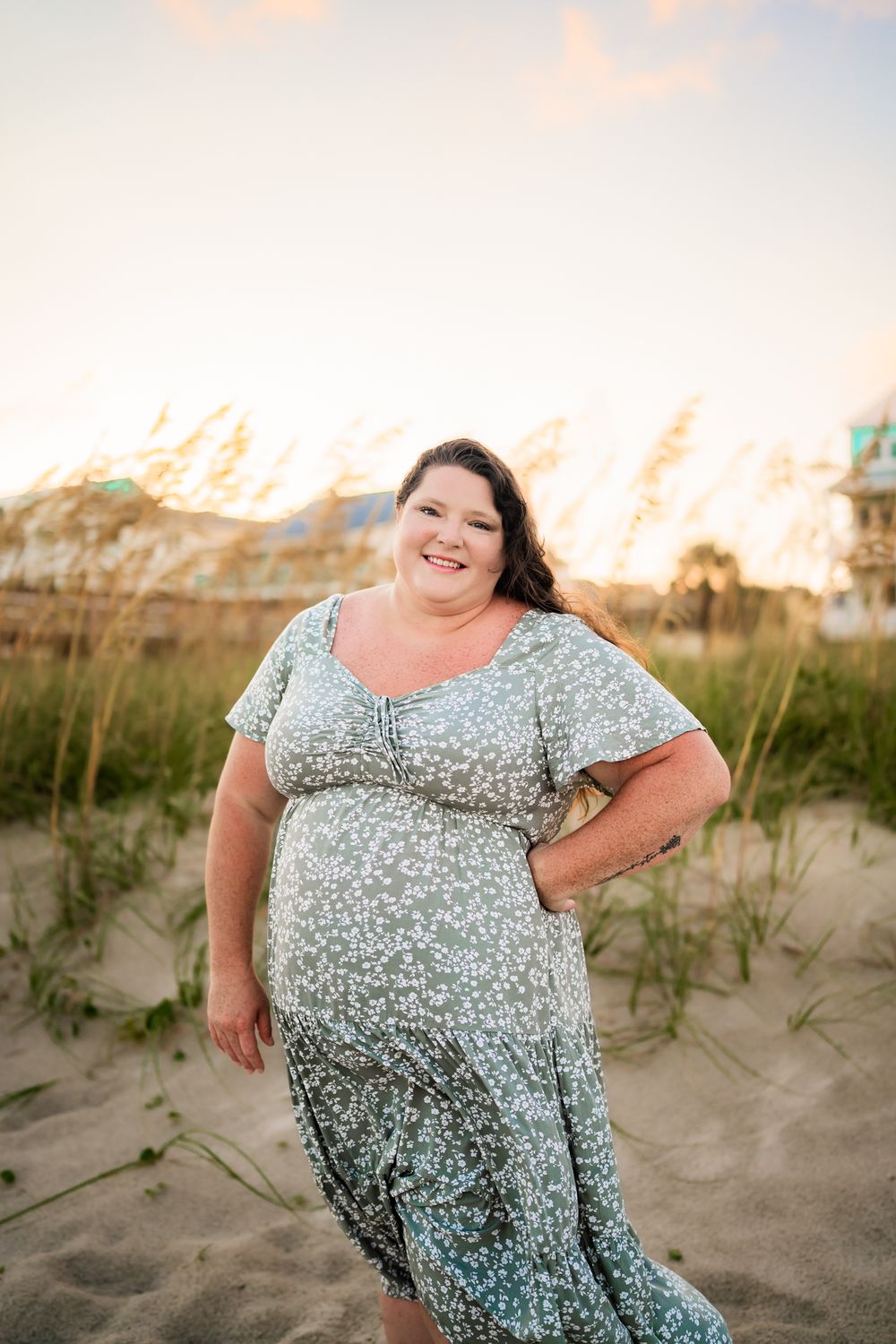 A person in a floral dress poses joyfully on sandy beach dunes during golden hour.