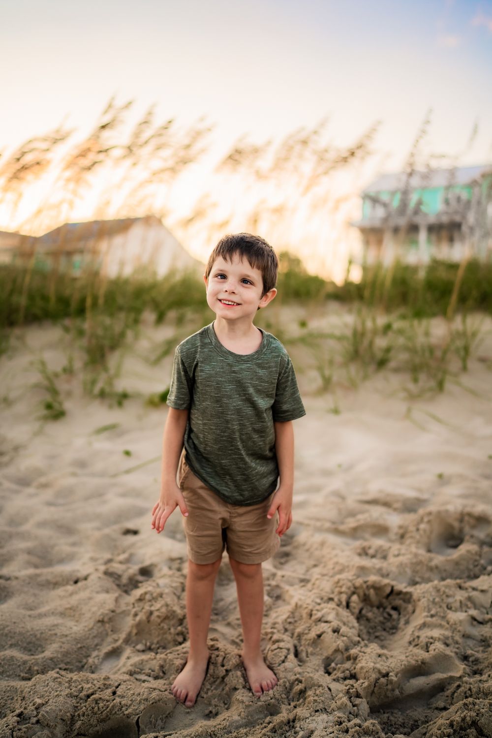 A child in olive green shirt stands happily on coastal sand dunes at sunset.