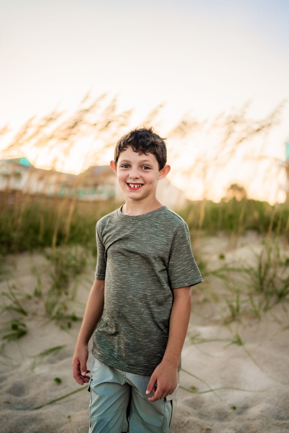 A young child in a green t-shirt stands on sandy dunes at sunset with beach grass in the background.