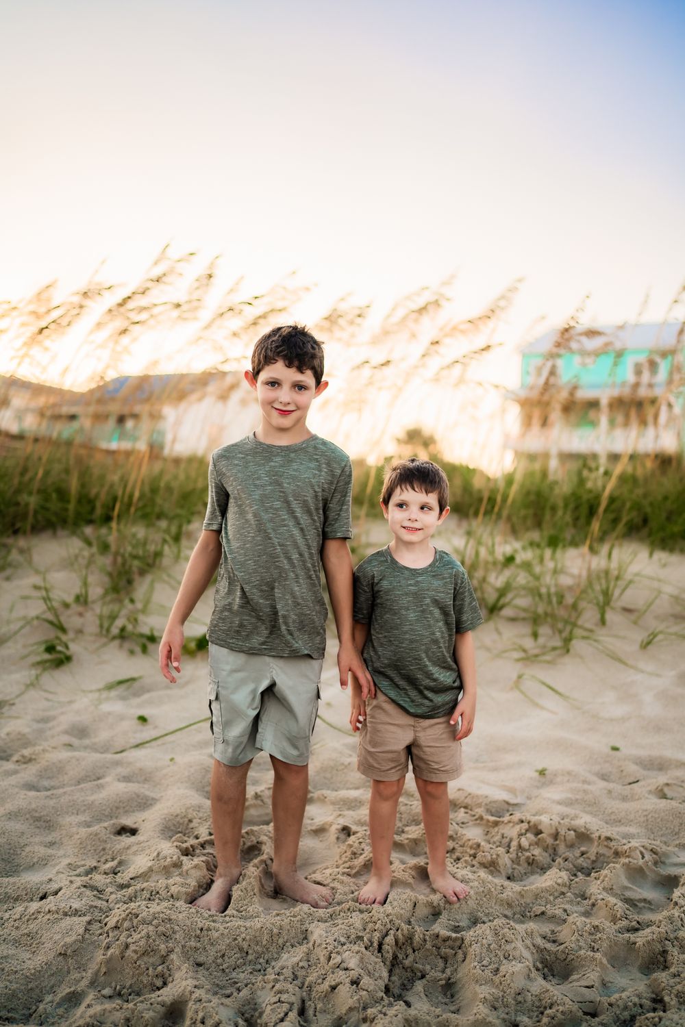 Two children stand together on a beach with sand dunes and beach grass at sunset creating a peaceful coastal scene.
