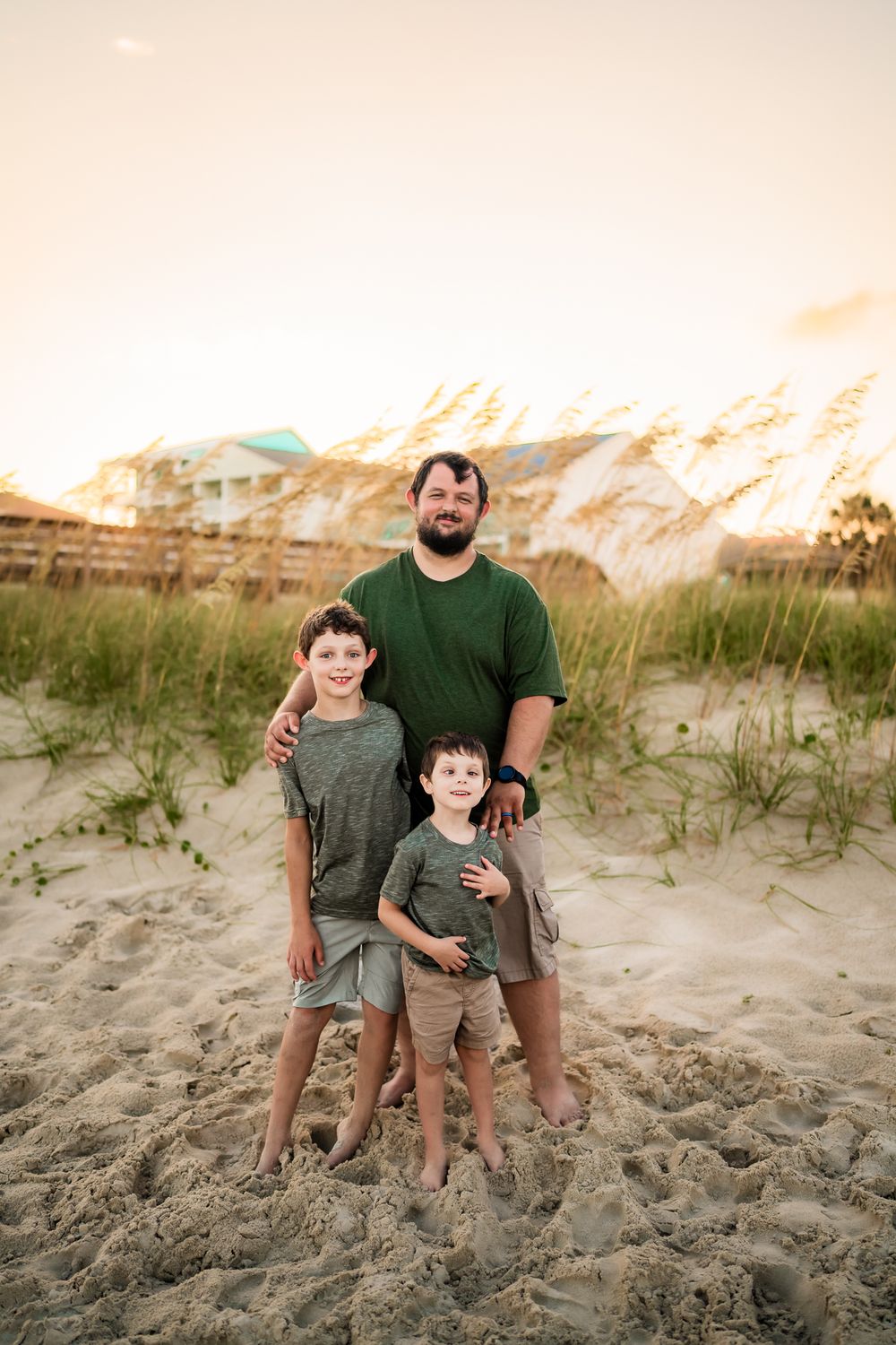 Three figures stand on sandy beach dunes at sunset, wearing casual clothing in earth tones.