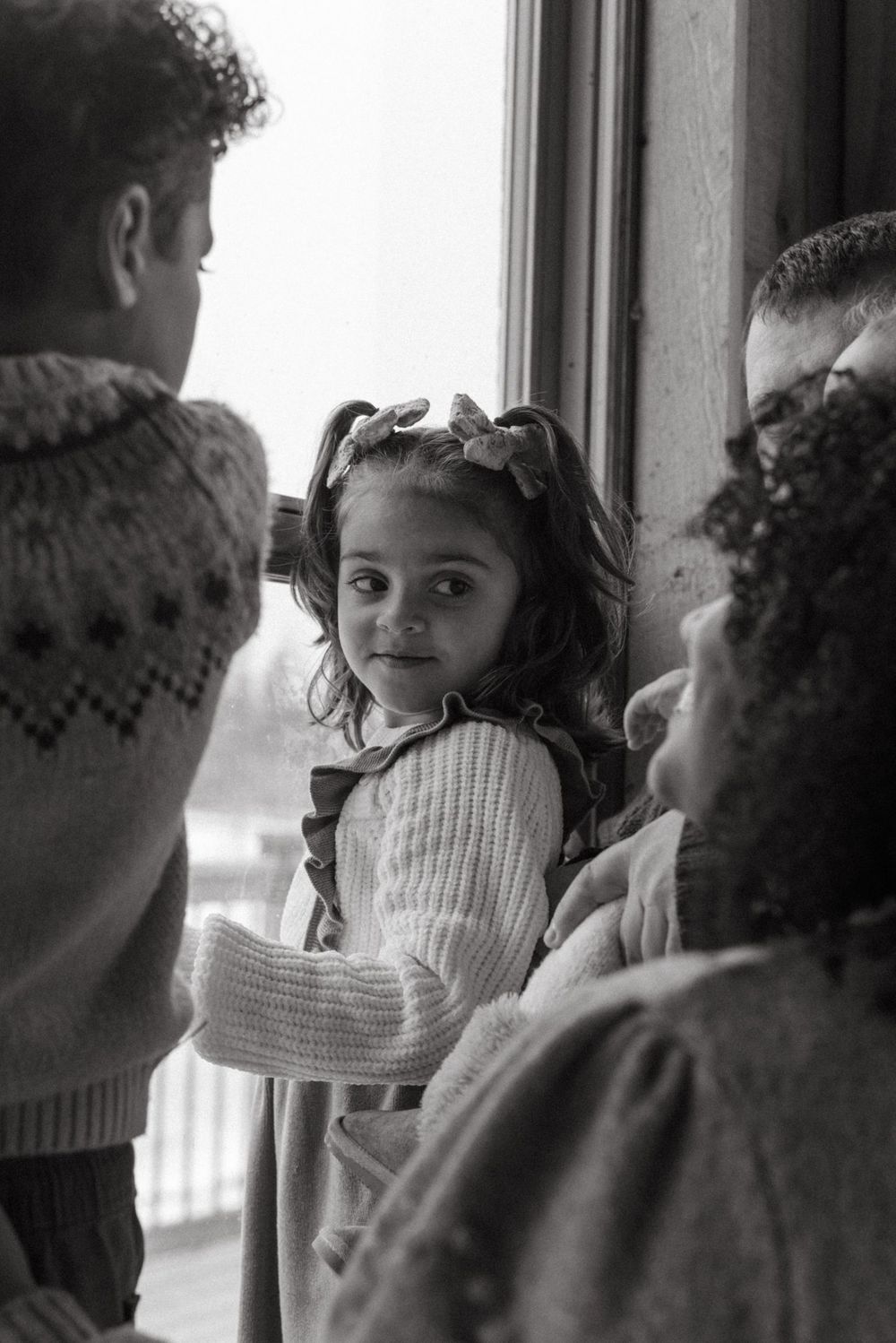 A candid black and white photograph of a young child in a white sweater looking at others by a window.
