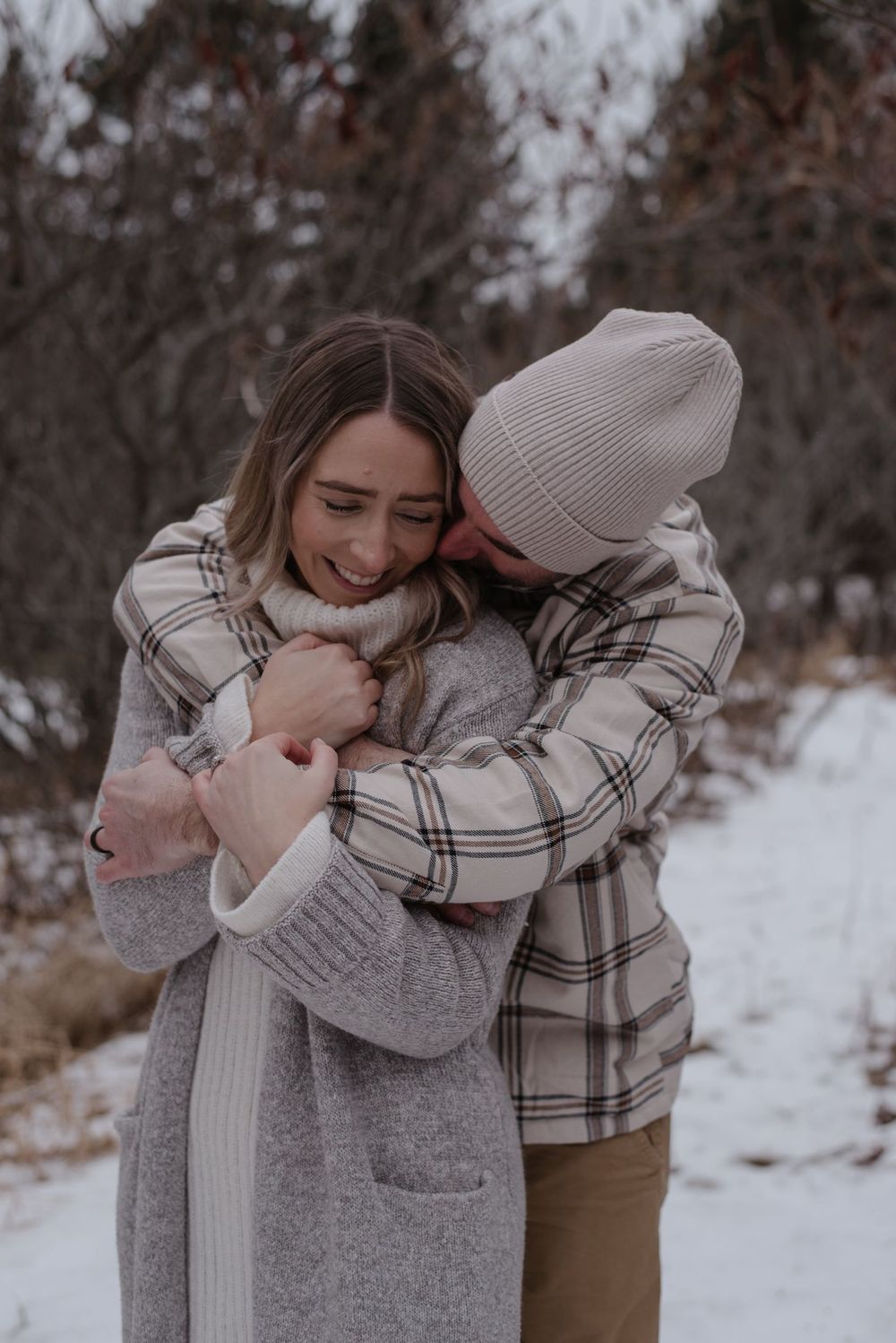 A couple embraces in a snowy winter setting, wearing cozy sweaters and plaid as they share an intimate moment outdoors.