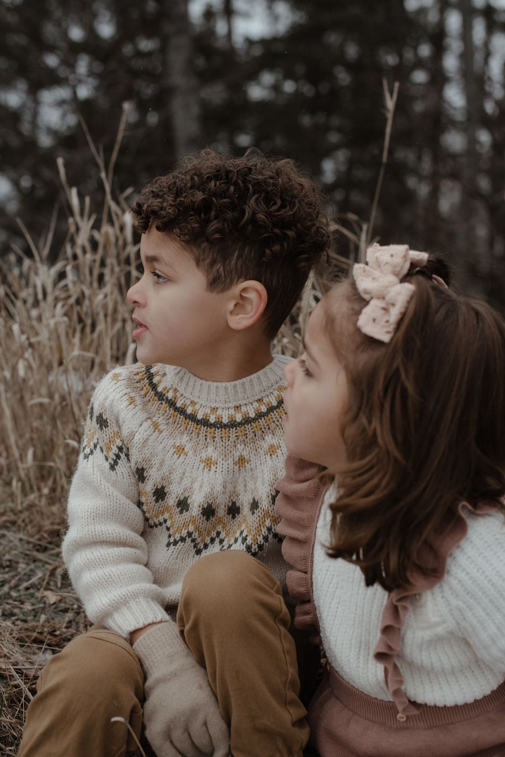 Two children in winter sweaters pose together against a backdrop of dried reeds in a nature setting.
