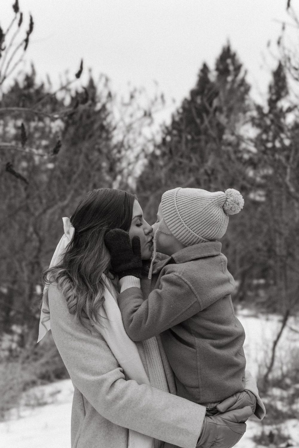 An intimate winter embrace between two people in warm clothing against a snowy evergreen forest backdrop.