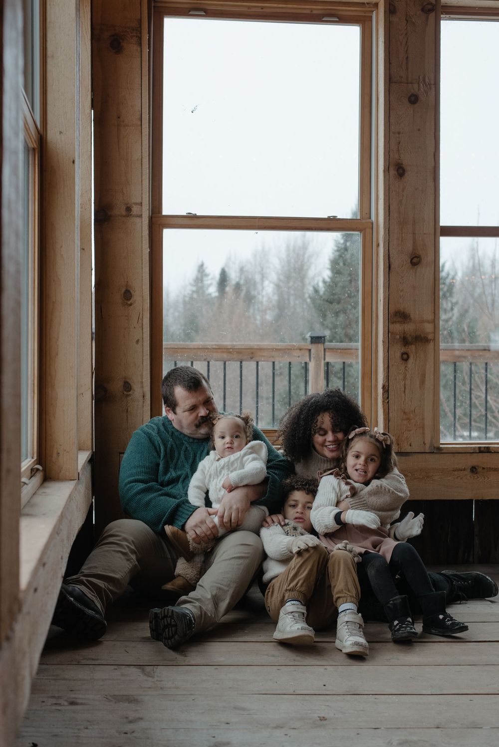 Family lounging together in sunlit room with wooden floors and large windows.