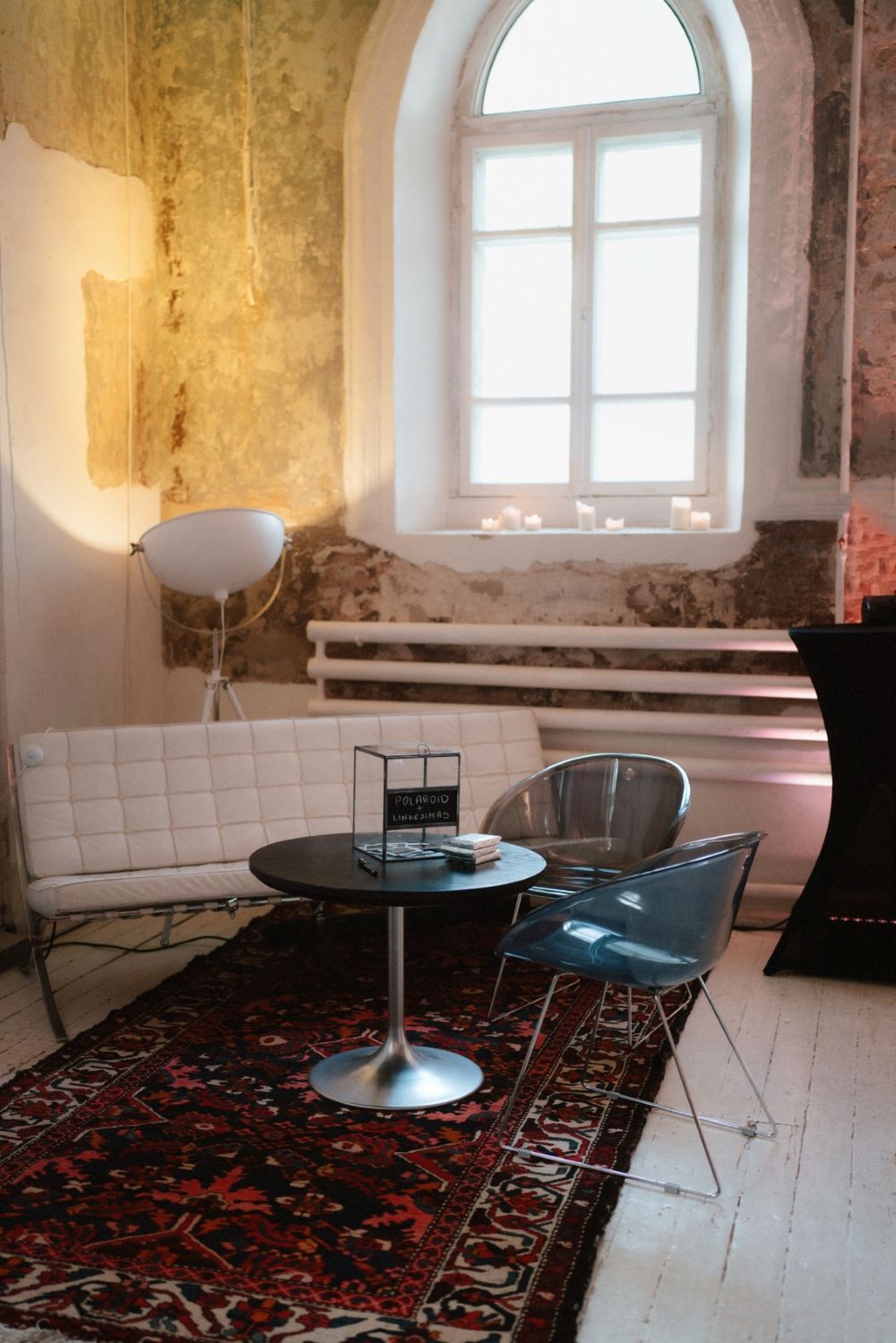 Modern lounge area with white leather bench, metal chairs and round table on ornate rug beneath arched window.