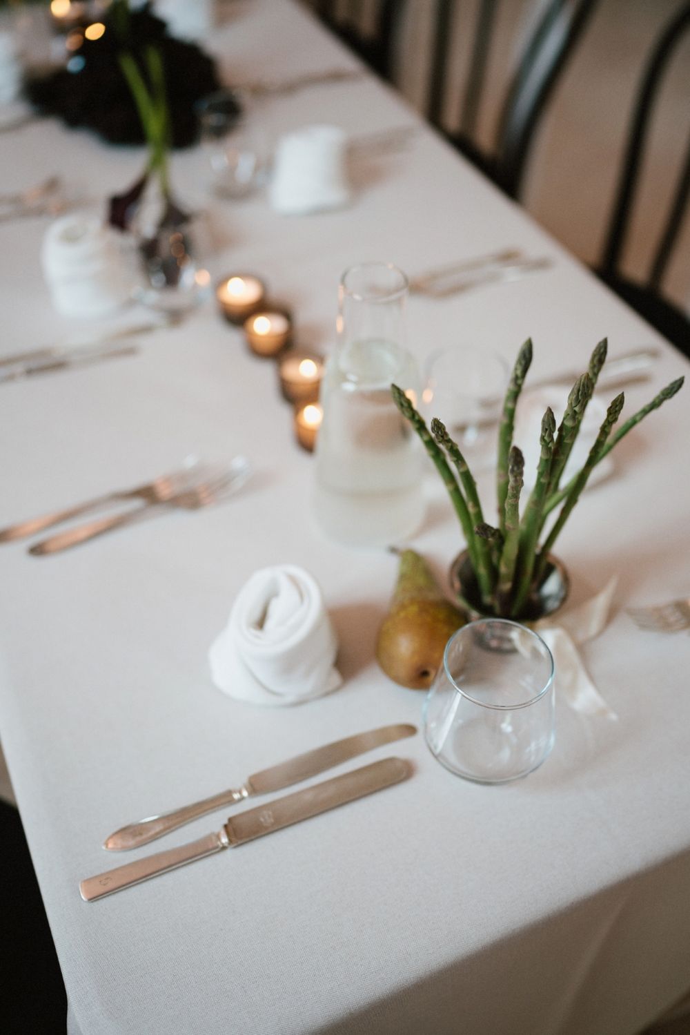 Minimalist table setting with asparagus in vase, candles, and white linens.