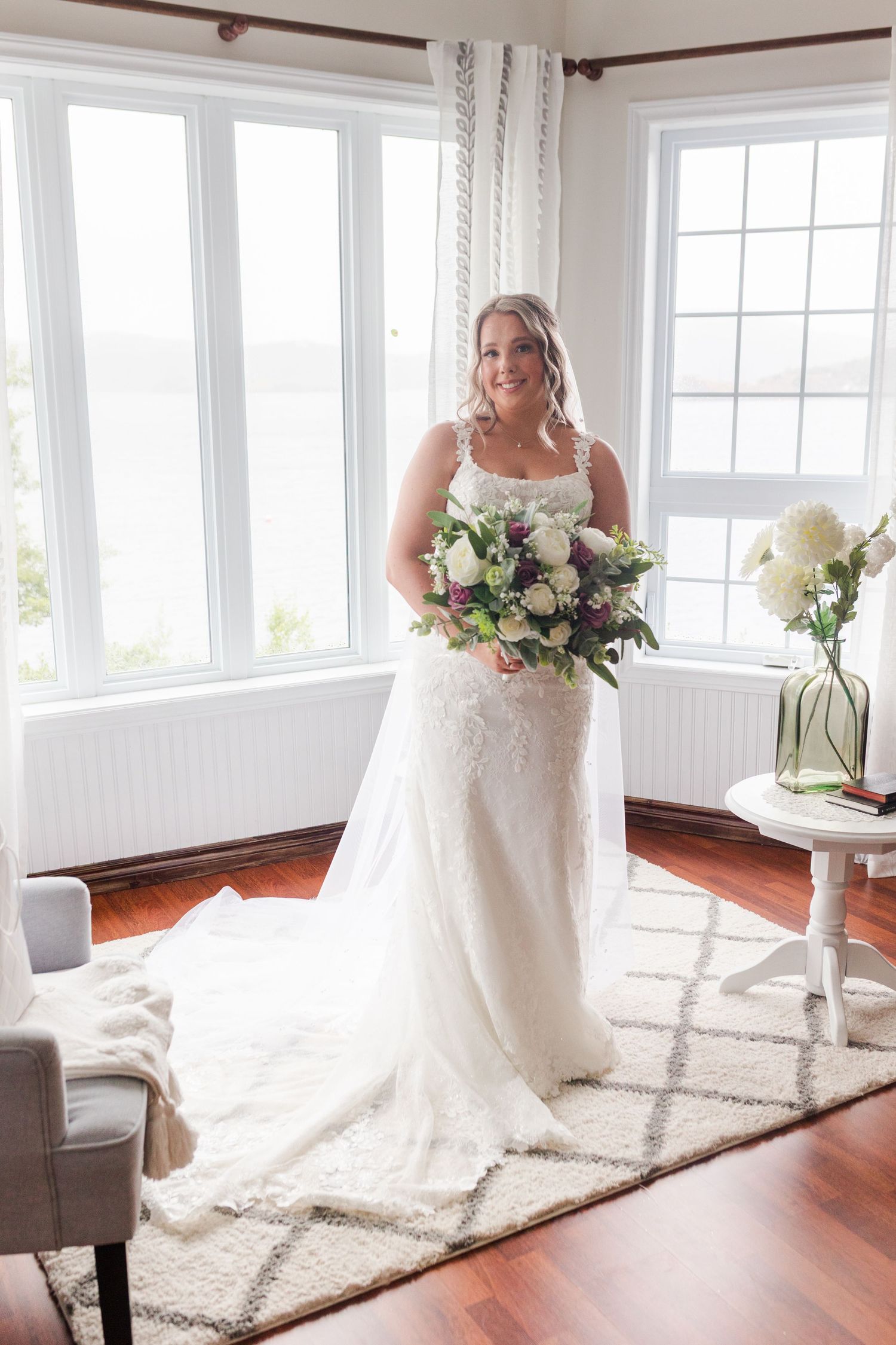 A bride in a flowing white wedding gown poses on a plush area rug in front of large windows in a bright sunlit room.