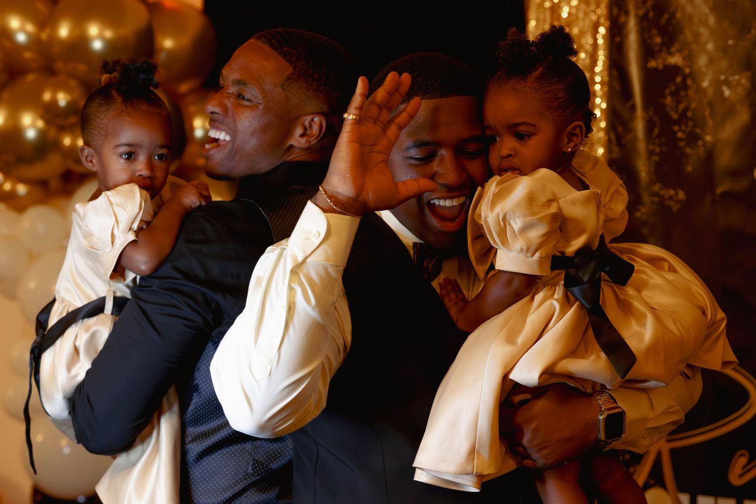Two men each holding a young child in formal attire at an elegant gold-themed celebration event.