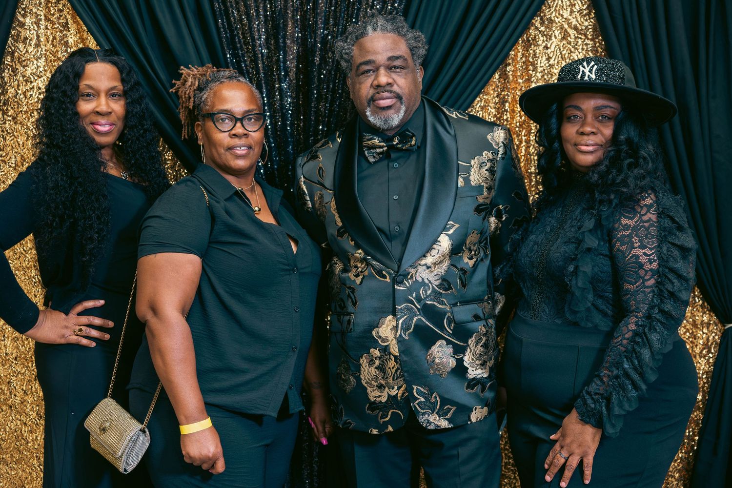 Four people dressed in elegant black attire pose before a gold sequin backdrop.