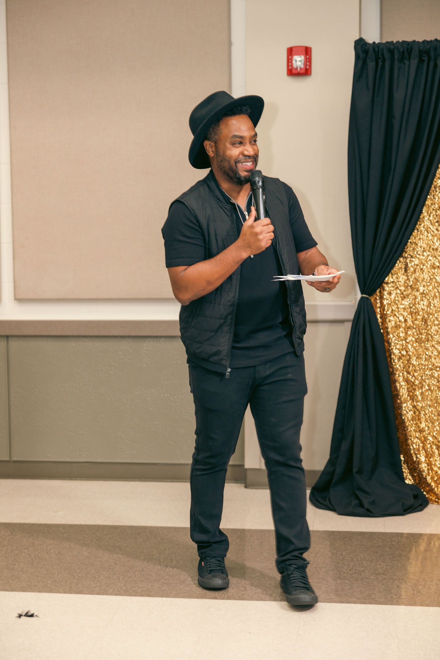 Stylish man in black outfit and hat speaks into microphone at a formal event.
