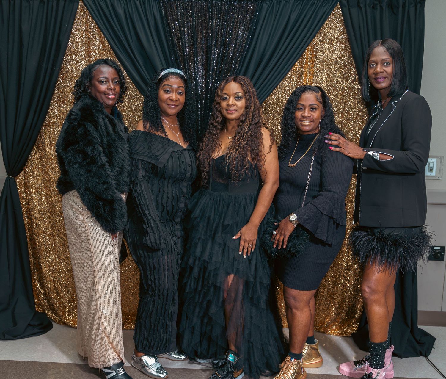 Five women in black outfits posing together in front of a gold sequin backdrop.