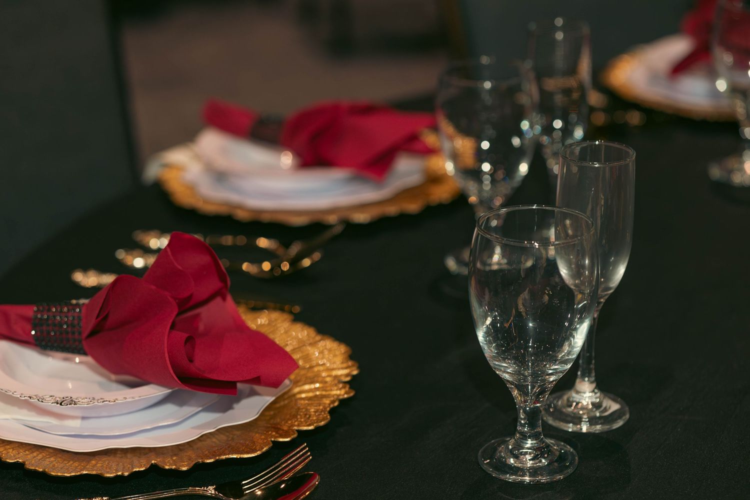 Elegant holiday table setting with red napkins, gold charger plates, and crystal glassware on black tablecloth.