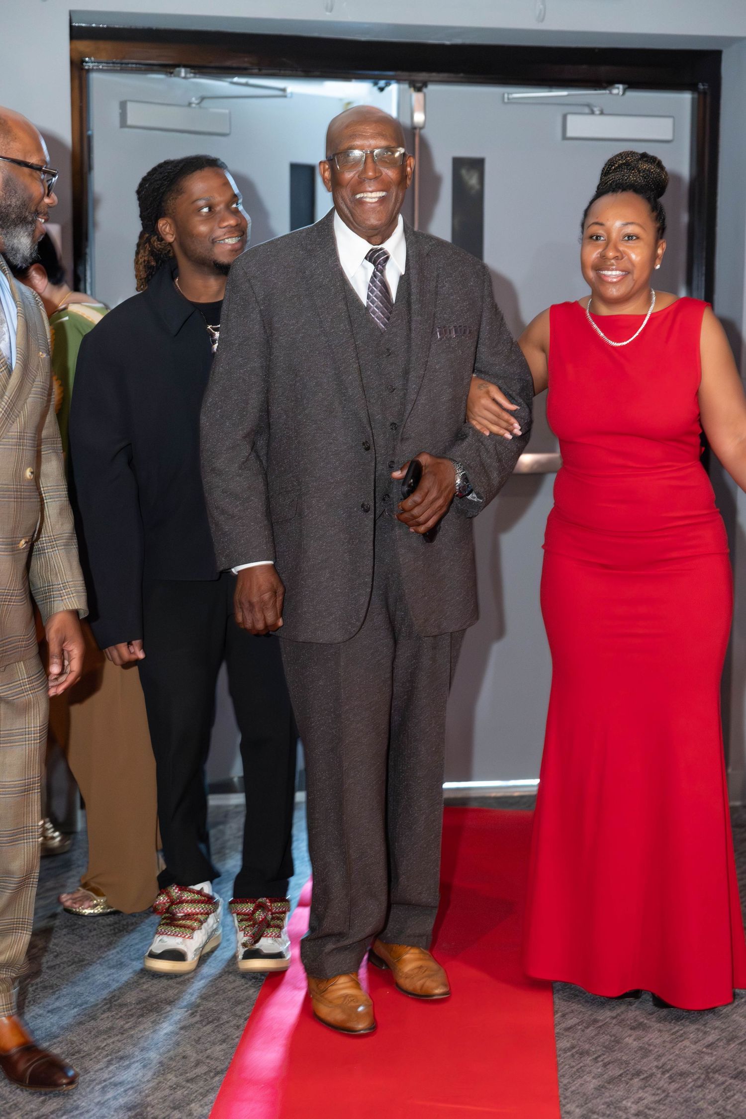 Elderly man in a gray suit escorted by a woman in red dress walking a red carpet.