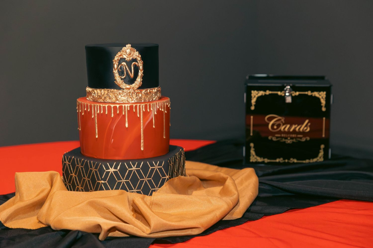 Elegant three-tier red and black birthday cake with gold drip decoration beside a cards box.