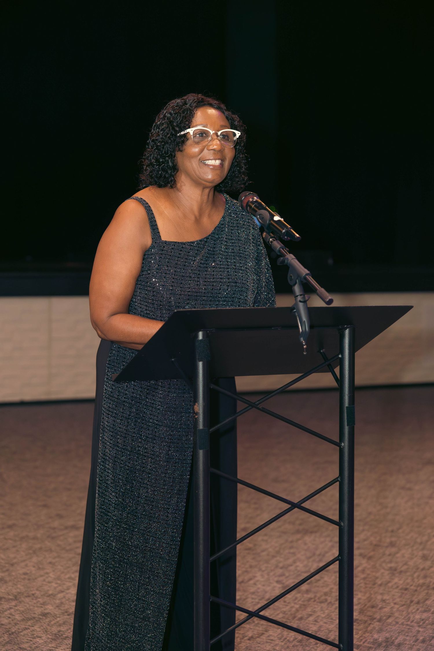 Woman in sparkly blue gown speaks at podium during formal event.