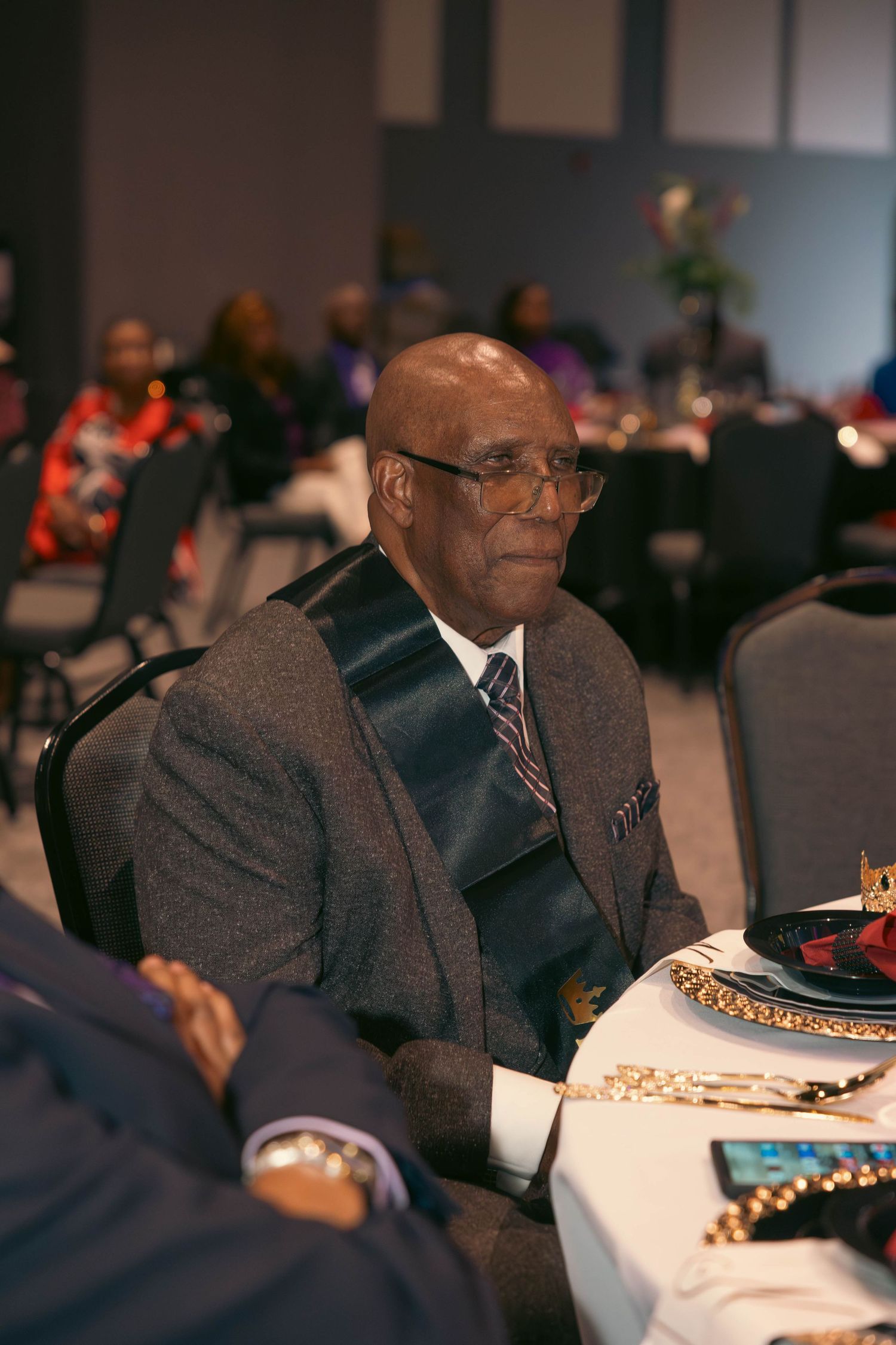 Elderly man in suit with sash sits attentively at formal banquet table.
